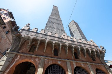 Low-angle view of the medieval Asinelli and leaning Garisenda Towers in brick and stone dominating central Bologna under a clear sky.