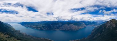 Dik orman yamaçları ve dramatik yaz bulutları altında engebeli uçurumlarla çerçevelenmiş derin bir dağ gölünün geniş hava manzarası..