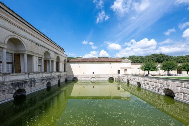 Mantua 'daki Rönesans Sarayı' nda, sıva kaplamalı perde ve taş köprünün yaz aylarında gökyüzünün altındaki yeşil hendeğe yansımasını görüyoruz..