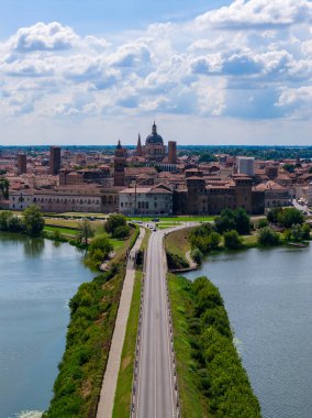 Aerial view of Mantuas medieval center and causeway between artificial lakes, highlighting brick towers, domes and Renaissance skyline in summer light.
