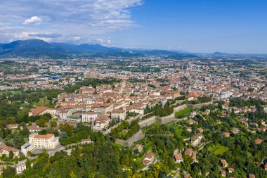 Bergamo 'nun yüksek hava manzarası terracotta çatılarını, yoğun kentsel kumaşı, yeşil tepeleri ve açık bir yaz gökyüzünün altındaki uzak Alpleri gösteriyor..