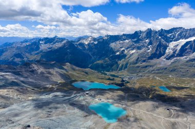 Valtournenche 'nin yukarısındaki kayalık bir buzul havzasında turkuaz Lago Goillet' in yüksek açılı görüntüsü, engebeli Pennine Alpleri ve yaz bulutları tarafından çerçevelenmiş..