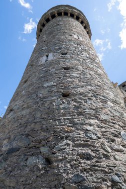 Low-angle view of the medieval stone keep of Fenis Castle in Valle dAosta, showing rough masonry walls and machicolations against a blue sky.