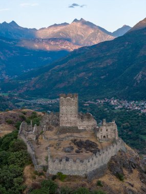 Günbatımında Aosta Vadisi 'ne bakan Ortaçağ Castello di Cly kalesi, Alp manzarasına hükmeden taş duvarlar ve kuleler.