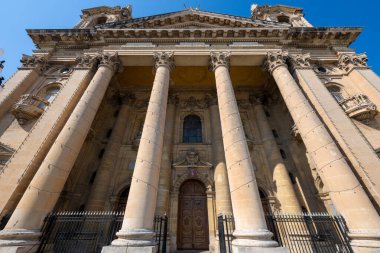 Valletta, Malta 'daki St. Publius Kilisesi' nin alçak açılı görüntüsü, yüksek Corinthian sütunları, ahşap kapı aralığı ve berrak mavi gökyüzü altındaki pedikürlü kireçtaşı Barok cephesini gösteriyor..