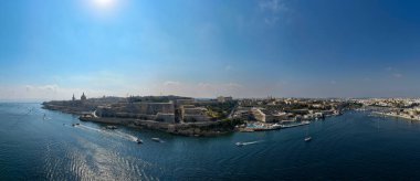 Vallettas St. Andrew Kalesi 'nin geniş panoramik manzarası ve Marsamxett Limanı üzerindeki tahkimatlar, açık bir yaz gökyüzü altında derin mavi Akdeniz sularında kayıklar..