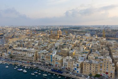 Grand Harbour boyunca uzanan Birgu (Vittoriosa) sahil manzarası yoğun kireçtaşı binalar, marina yatları ve gün batımında bulutlu bir Akdeniz gökyüzünü gösteriyor..