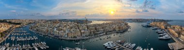Gün batımında Maltas Grand Harbour 'daki limanın geniş panoramik manzarası, yat, kireçtaşı binalar ve su üzerindeki tahkimatlar.