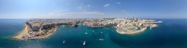 Sliema ve St Julians kıyı şeridinin geniş bir manzarası, Malta, turkuaz Akdeniz Körfezi, marinalar, kayalık kıyılar ve açık yaz gökyüzü altında demirlemiş yelkenliler.