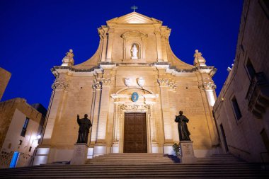 Victoria Katedrali 'nin (Rabat) aydınlanmış Barok cephesi, Gozo, derin mavi gökyüzünün altında iki heykel ve basamakla.