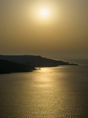 Ramla Bay Beach, Nadur, Malta 'da kıyı şeridi boyunca yürüyen bir insanın silueti, sakin deniz suyunda düşük güneş ve altın yansımalarıyla.