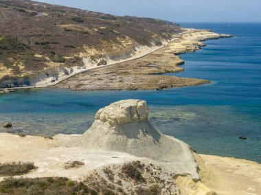 Zebbug, Malta yakınlarındaki Xwejni Kayası 'nda kayalık bir sahil şeridi, soluk kireçtaşı çıkıntısı, kıyı yolu ve berrak yaz ışığı altında sakin mavi Akdeniz suyu.