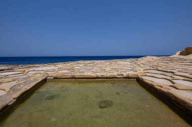 Kireç taşına oyulmuş dikdörtgen deniz suyu tuzu tavası ve açık mavi gökyüzünün altında Akdeniz kıyılarına uzanan geleneksel tuz tavaları..