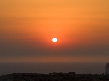 Turuncu gün batımı, Akdeniz ufkunun üzerinde güneş diski, puslu eğimli gökyüzü ve sakin su, Sannat, Gozo, Malta yakınlarındaki Cenc Kayalıklarından görülüyor..