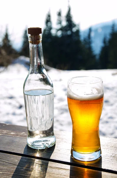 Μπύρα και μπουκάλι σκληρό ποτό σε ξύλινο τραπέζι στα βουνά του χειμώνα — Φωτογραφία Αρχείου
