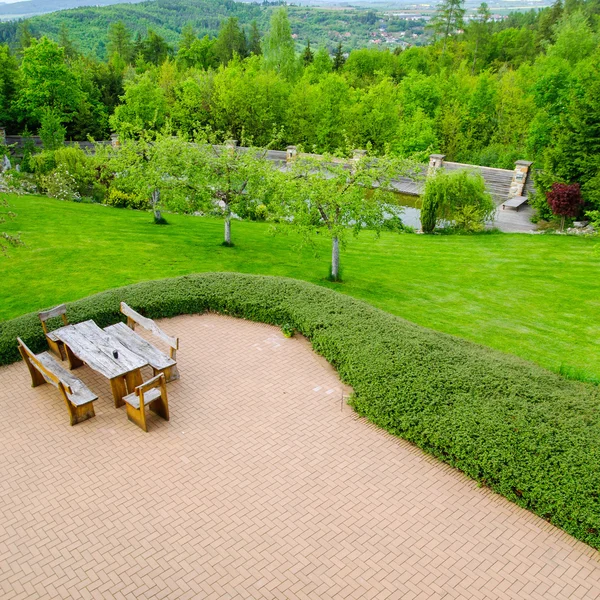 Tegel stenlagd uteplats med bekväma trädgårdsmöbler med rustik fåtöljer och en pall omgivet av garden, hög vinkel Visa — Stockfoto