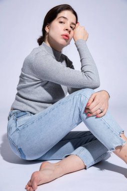 Genç bir kadın, elleri griye yakın, kot pantolon içinde oturuyor.