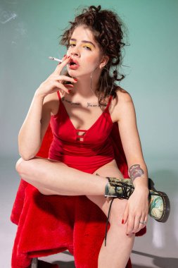 Sigara içen beyaz manken kırmızı elbiseli, yeşil renkli sandalyede oturuyor.