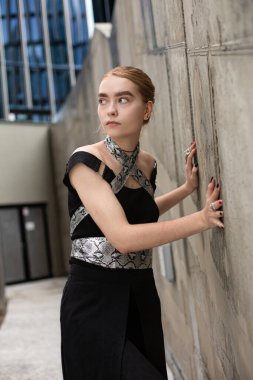 Siyah elbiseli beyaz kadın duvarın yanında duruyor.
