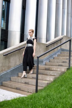 Siyah elbiseli beyaz kız merdivenlerden aşağı sütunların yanına iniyor.