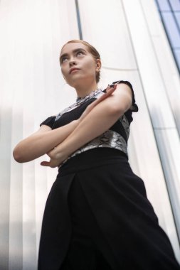 Siyah elbiseli, ciddi sarışın kadın. Metal paneller arkasında.