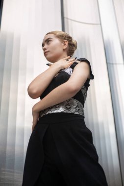 Siyah elbiseli, ciddi sarışın kadın. Metal paneller arkasında.