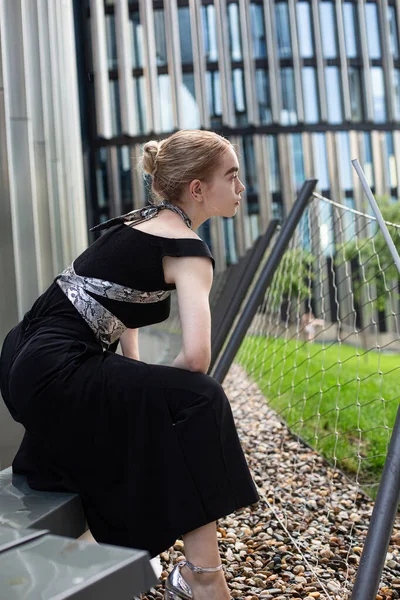 Siyah elbiseli beyaz kadın şehir yolunda oturuyor.