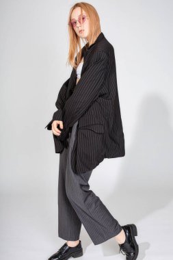 Güneş gözlüklü ve ceketli Avrupalı genç bir kadın gri üzerinde tek başına duruyor.