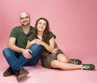 Mutlu beyaz çift pembe renkte izole olmuş gülümsüyor ve yerde oturuyor.