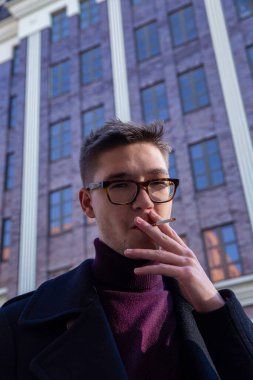 Beyaz Avrupalı genç adam arkasında şehir binası olan bir sigarayla
