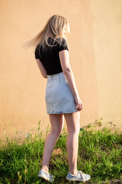 White young woman in skirt with blond hair near yellow wall and green grass