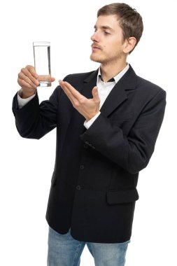 Genç yakışıklı, uzun boylu, zayıf, kahverengi saçlı, beyaz arka planda siyah ceketli bir bardak su gösteren beyaz bir adam.