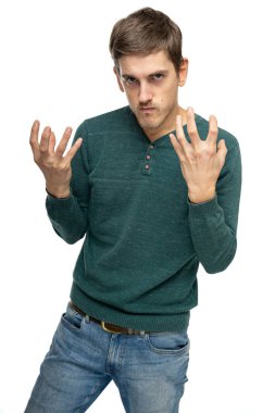 Genç yakışıklı, uzun boylu, zayıf, kahverengi saçlı, beyaz arka planda izole edilmiş mavi kot pantolonlu, koyu yeşil süveter içinde öfkeli görünüyor.