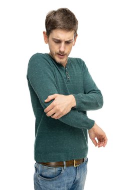 Genç yakışıklı, uzun boylu, zayıf, kahverengi saçlı, dirseğini koyu yeşil kazaklı, beyaz arka planda izole edilmiş kot pantolonlu bir adam tutuyor.