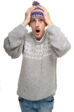 Genç yakışıklı, uzun boylu, zayıf, kahverengi saçlı beyaz adam beyaz arka planda izole edilmiş renkli kış şapkalı gri kazakla şaşırmış görünüyor.