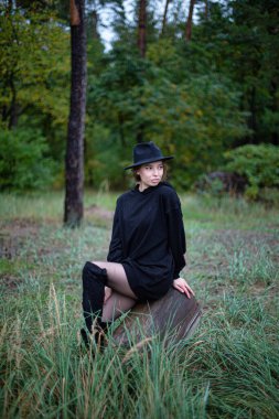 Siyah elbiseli, arkasında orman olan beyaz bir kadın.