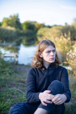 Uzun saçlı beyaz Avrupalı genç adam gölün arkasında oturuyor.