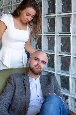 Beyaz, genç, evli, pencereli bir çift bir adam oturuyor ve arkasında bir kadın duruyor.