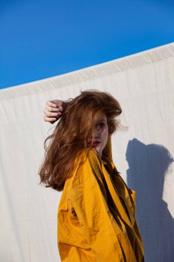 Duvarın yanında parlak makyajlı, arkasında açık gökyüzü olan genç beyaz kız.