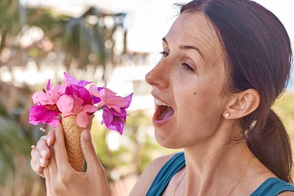 Menina de cabelos castanhos tentando comer sorvete com flores na frente de folhas de palmeira em tempo ensolarado. Conceito de publicidade de férias de verão. Verão flores surreais conceito criativo e moderno. — Fotografia de Stock