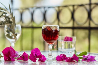 Zarif, parlak bir kadeh kırmızı şarap, kadehte şarap sıçraması. Arka planda yaz kokteylleri. Dekoratif pembe çiçekler ve bir bardak zeytin ağacı dalı. Arındırmanın Kabartması