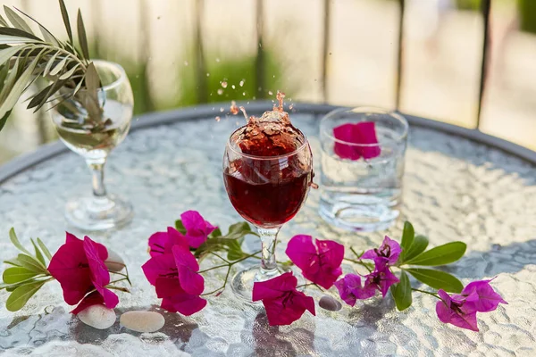 Elegant and colorful explosion of red wine. Summer cocktails on the background. Decorative pink flowers and a branch of an olive tree in a glass. Glyph of Purification. Copy space.