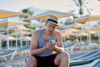 Sahilde, deniz kenarında bir plaj şemsiyesinin altında telefonu olan genç bir adam. Tatil konsepti, arkadaşlarla veya serbest çalışanlarla sohbet, uzaktan çalışma, webinar, iletişim, öğrenme konsepti