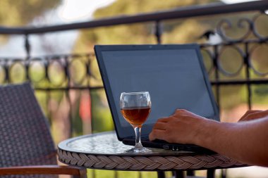 Dizüstü bilgisayarı ve bir bardak şarabı olan biri güneşli bir günde otel balkonunda serbest çalışır. Kendini soyutlama, ev konseptinde çalışma. Çevrim içi iş