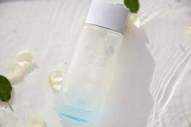 Parfüm bardağı şeffaf şişe, beyaz gül yaprakları ve yaprakları ile suda taklit edilir. Yüksek açılı gölgeler. Üst Manzara. Mesaj için yer. Yüksek kalite fotoğraf