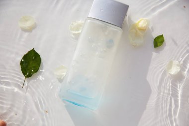 Parfüm bardağı şeffaf şişe, beyaz gül yaprakları ve yaprakları ile suda taklit edilir. Yüksek açılı gölgeler. Üst Manzara. Mesaj için yer. Yüksek kalite fotoğraf