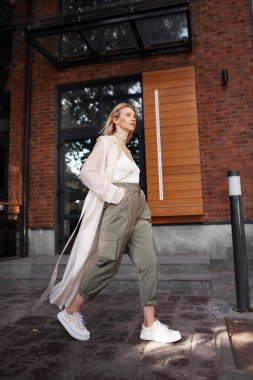 Kadın Modeli Yürüyen Şehir Sokağı. Moda Kadını Blogcusu Moda Giyen İlkbahar ya da Sonbahar Giysileri (bej renkli trençkot, büyük beden haki pantolon, aksesuar) açık havada. Trend Sokağı Kıyafeti