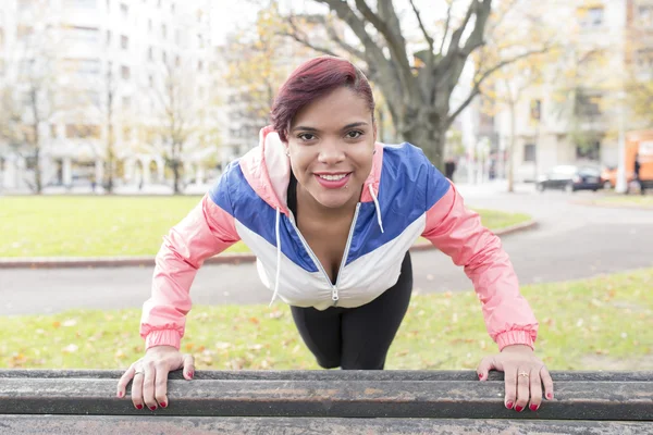 Mujer joven sonriente flexiones en el banco de madera en el parque . — Foto de Stock