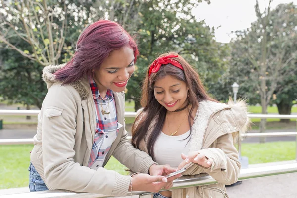 Dos amigas sonrientes compartiendo redes sociales en un teléfono inteligente . — Foto de Stock