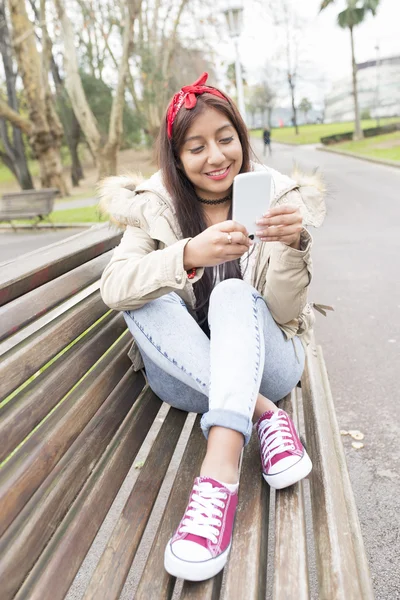 Mujer sonriente sentada en el banco con teléfono inteligente . —  Fotos de Stock